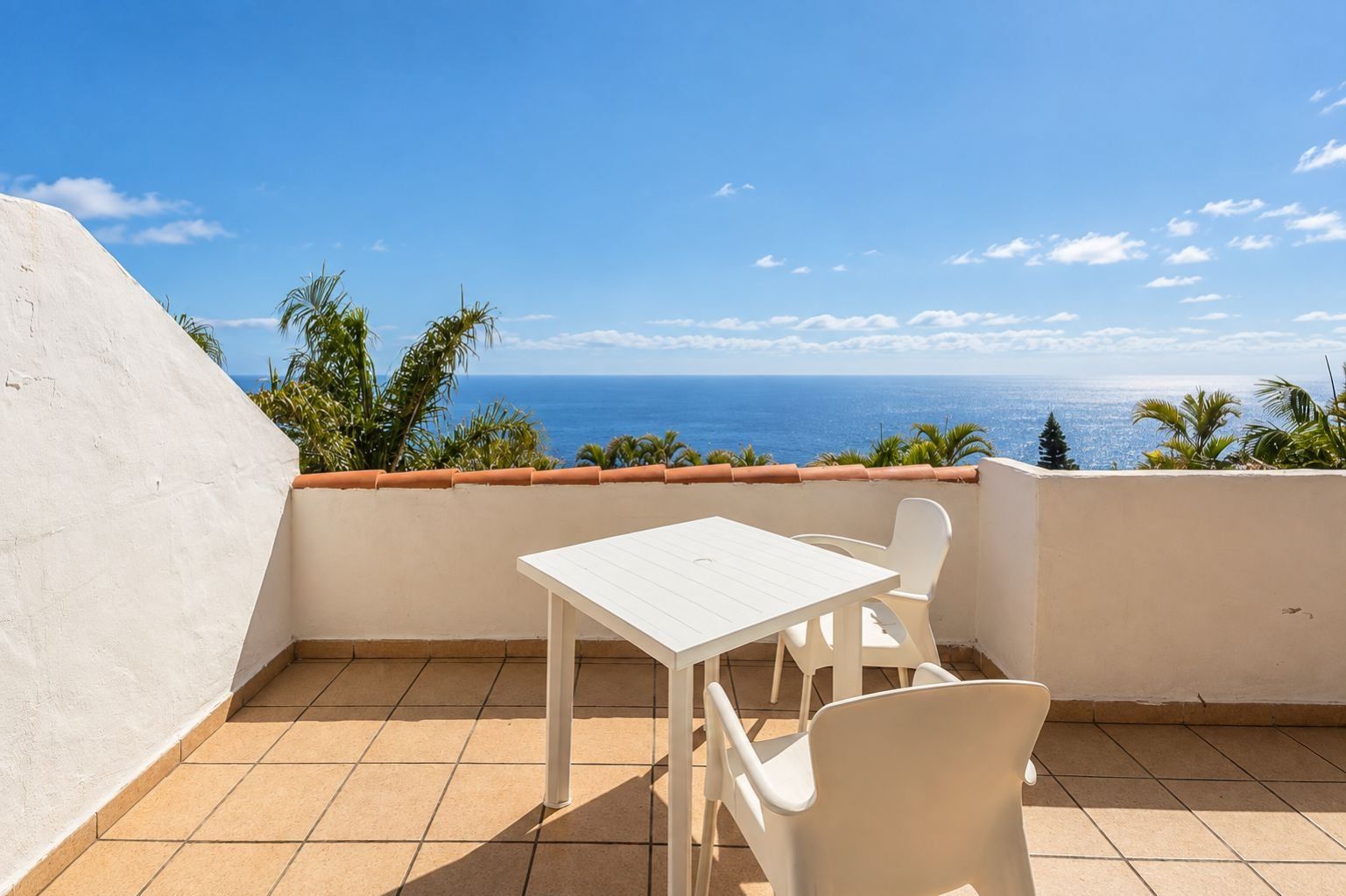 Een balkon met een witte tafel en stoelen, een betegelde vloer en uitzicht op de oceaan.