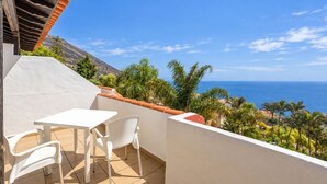 Un balcó amb taula i cadires, amb vista a un paisatge costaner de palmeres i mar.