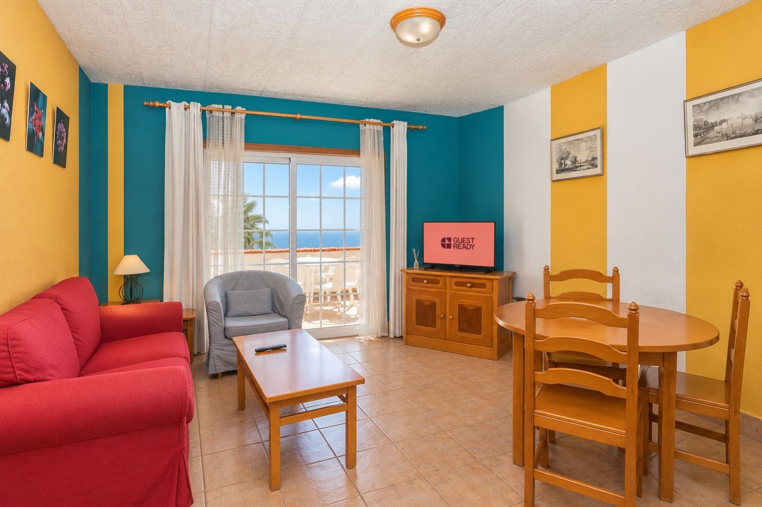 Una habitación con un sofá rojo, un sillón gris, una mesa de madera, un televisor sobre un mueble de madera y una ventana con cortinas.
