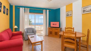 Una habitación con un sofá rojo, un sillón gris, una mesa de madera, un televisor sobre un mueble de madera y una ventana con cortinas.
