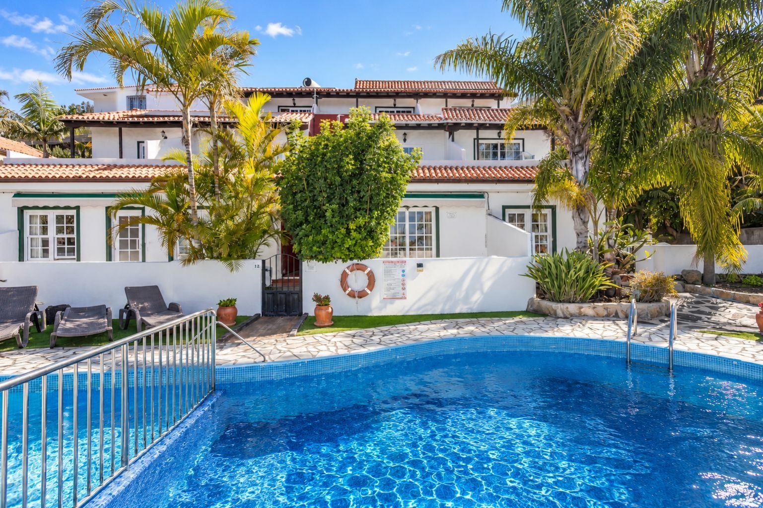 Una piscina con borde de azulejos, rodeada de sillones de descanso, palmeras y un edificio con techo de tejas.