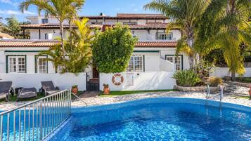Una piscina con borde de azulejos, rodeada de sillones de descanso, palmeras y un edificio con techo de tejas.
