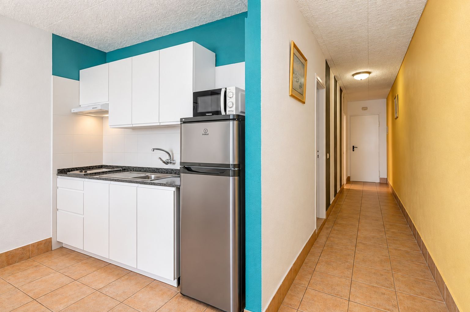 Una cocina compacta con muebles blancos, un refrigerador de acero inoxidable y un microondas.