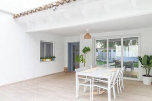 Een ruime kamer met een witte tafel en stoelen, een groot raam en een deur naar een buitenruimte.