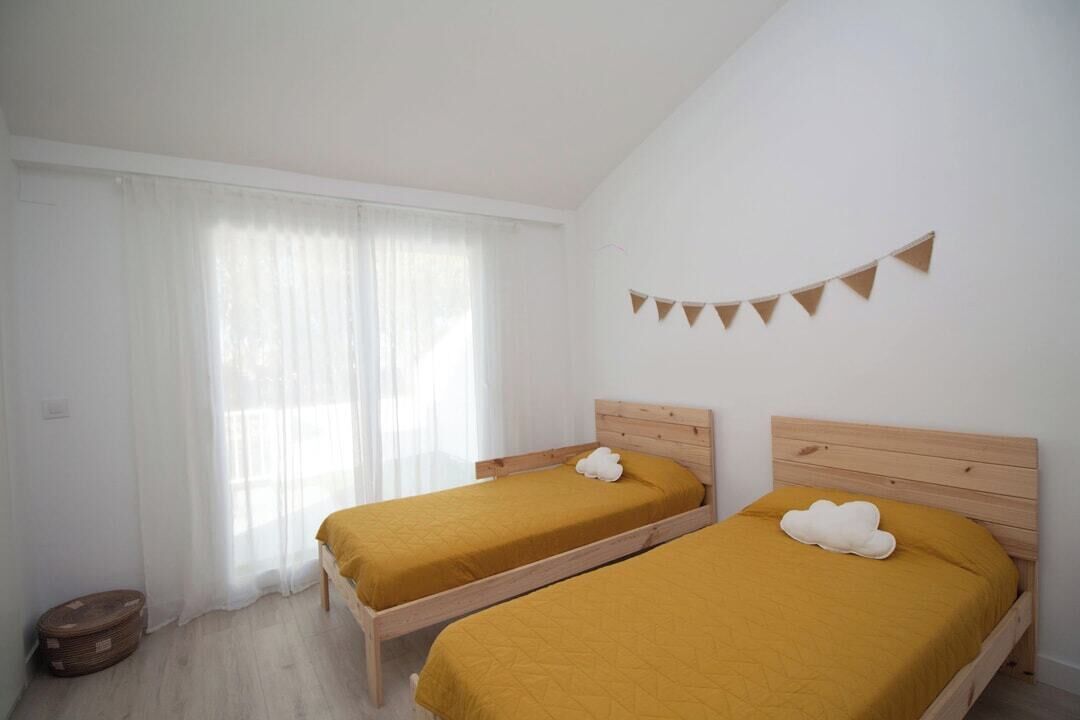 A room with two beds, a window with sheer curtains, a small round table, and a wall-mounted bunting decoration.