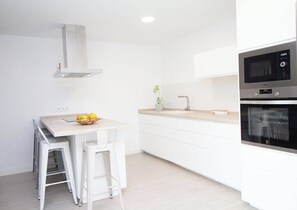 Eine moderne Küche mit weißen Schränken, einer Edelstahl-Dunstabzugshaube, einer Mikrowelle und einem Backofen. In der Mitte steht eine kleine Kochinsel mit Hockern und einer Obstschale.