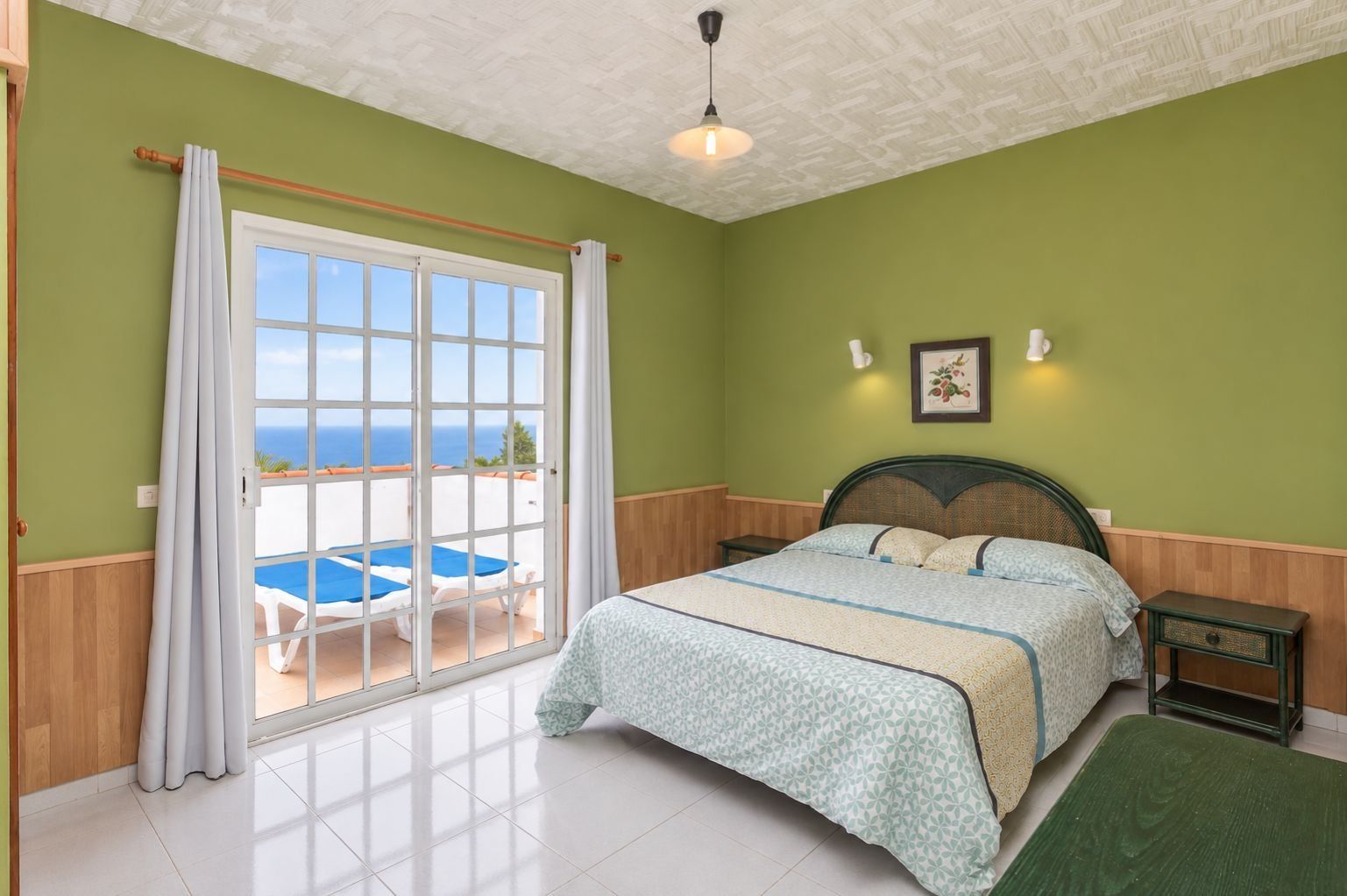 A bedroom with a bed, a nightstand, a green wall, a window with curtains, and a view of the sea.