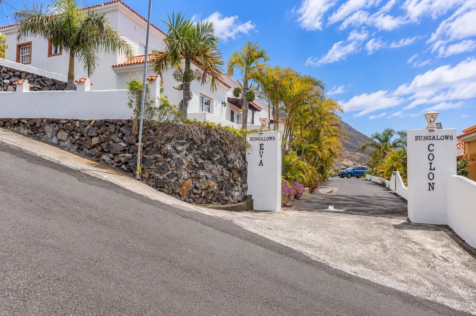 Una entrada protegida por una reja a una propiedad llamada 'BUNGALOWS EVA', con un muro de piedra y palmeras.