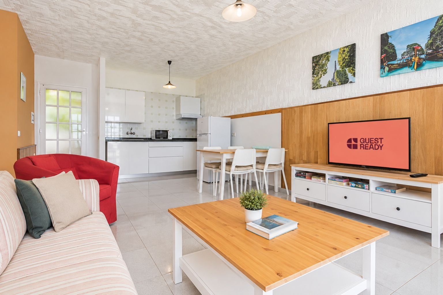 Ein modernes Wohnzimmer mit einer Couch, einem Couchtisch mit Buch und Pflanze, einem Fernseher, der 'GUEST READY' anzeigt, und einem Essbereich mit Stühlen und einem Tisch.