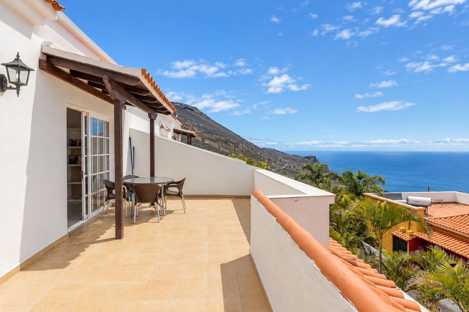 A balcony with a table and chairs, overlooking a coastal landscape with a view of the sea and hills.