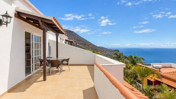 A balcony with a table and chairs, overlooking a coastal landscape with a view of the sea and hills.