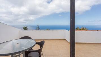 A balcony with a glass table and chairs, overlooking a coastal view.