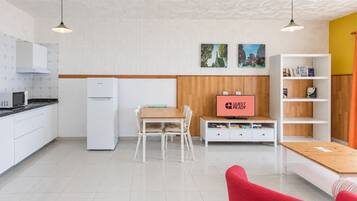 Eine moderne Küche mit weißen Schränken, einer Mikrowelle und einem Kühlschrank. Es gibt eine Essecke mit einem Holztisch und Stühlen, und ein Fernseher zeigt 'GUEST READY'.