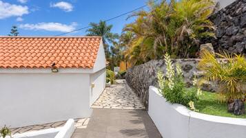 Un sendero estrecho que conduce a un edificio con techo de tejas, rodeado de abundante vegetación y un muro de piedra.