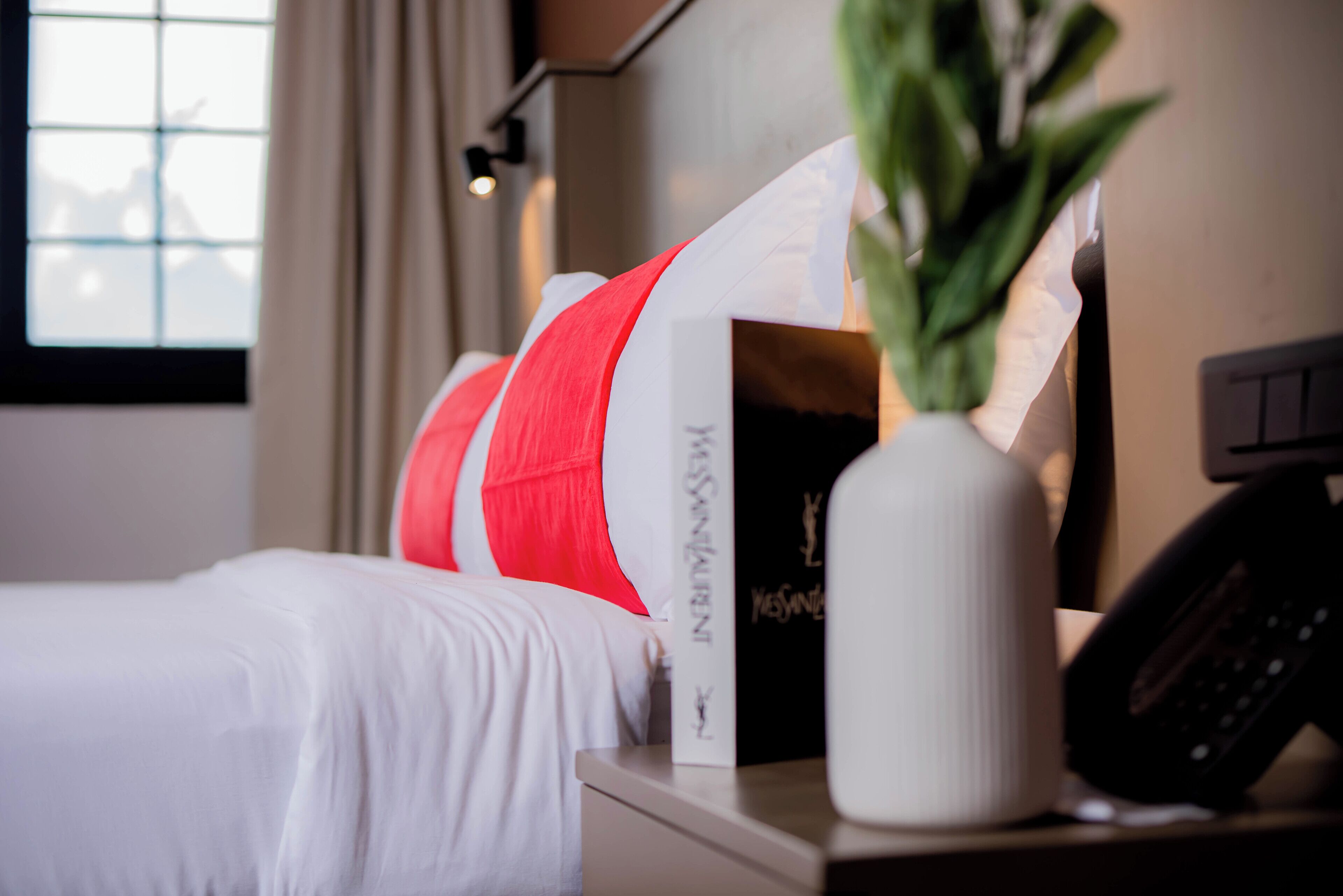 A neatly made bed with white bedding, a bedside table with a white vase and a black object, and a window with curtains.