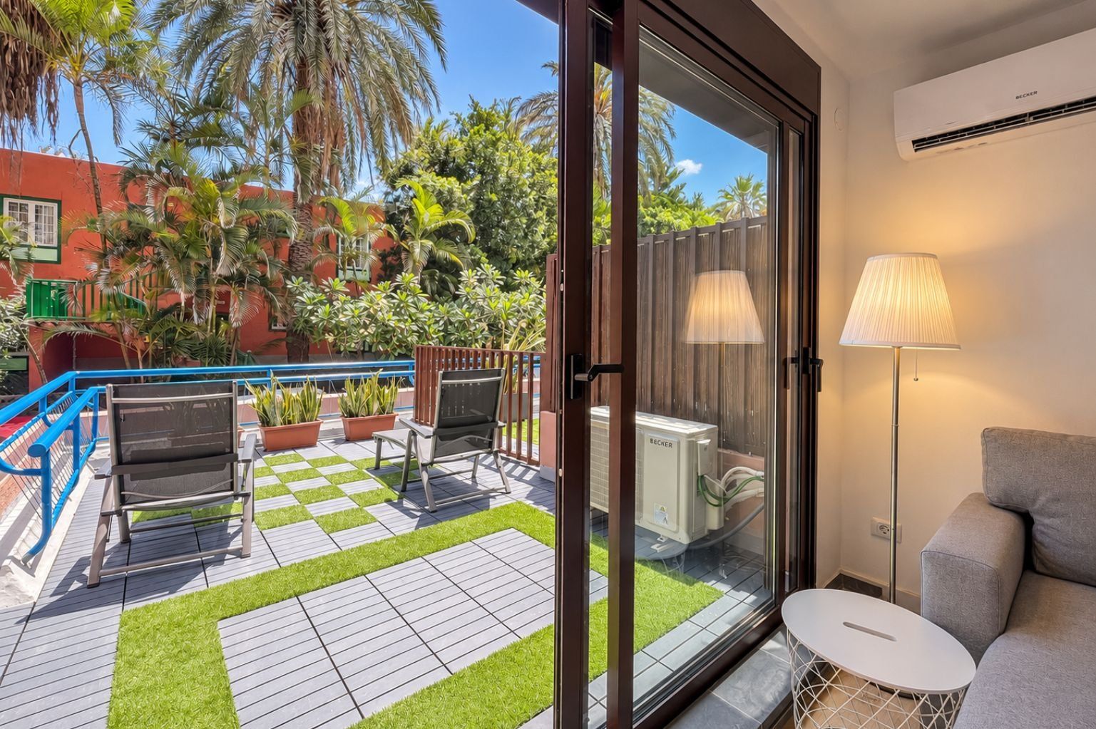 Una habitación con puerta de vidrio corredera que da a un patio con piso a cuadros, mobiliario exterior y plantas en macetas.