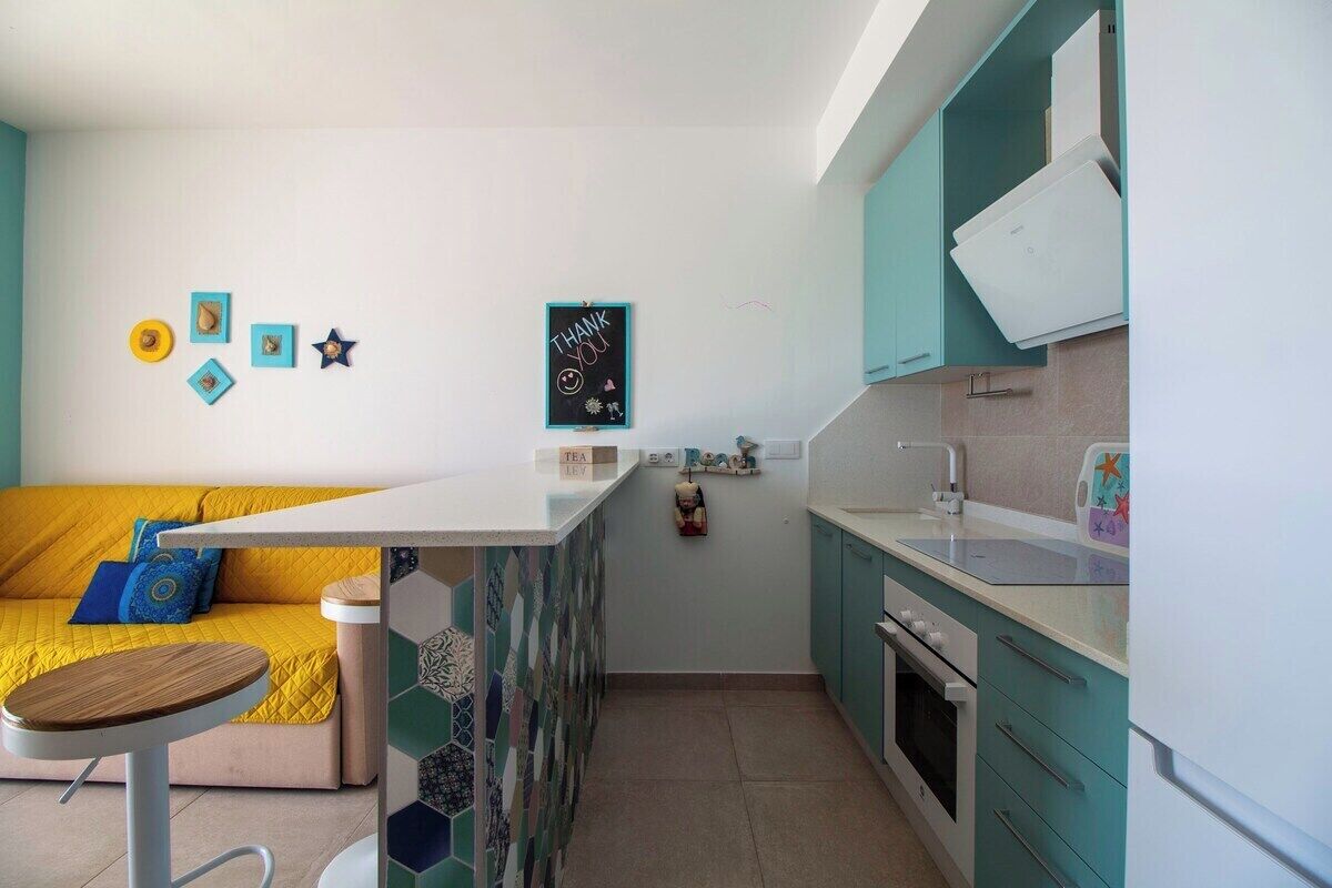 A compact kitchen with teal cabinets, a white countertop, and a small dining area with a yellow sofa.