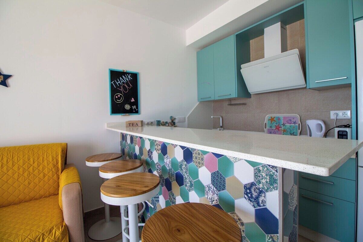 A compact kitchen with teal cabinets, a white countertop, and a colorful hexagonal backsplash. There are three stools and a yellow sofa.