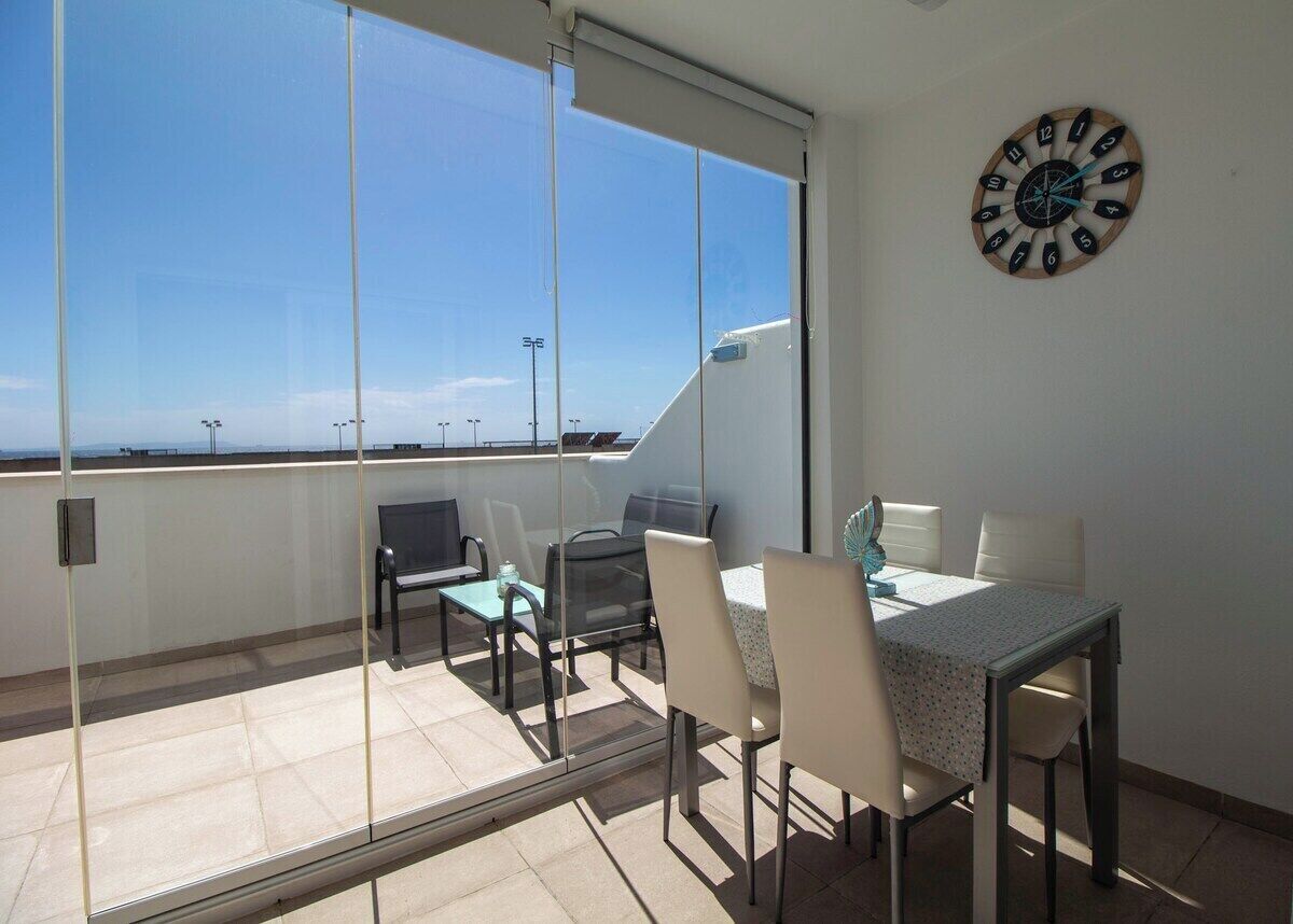 Een balkon met een tafel en stoelen, een klok aan de muur, en een helder uitzicht op de lucht.