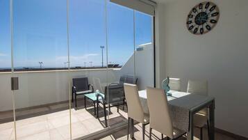 Een balkon met een tafel en stoelen, een klok aan de muur, en een helder uitzicht op de lucht.