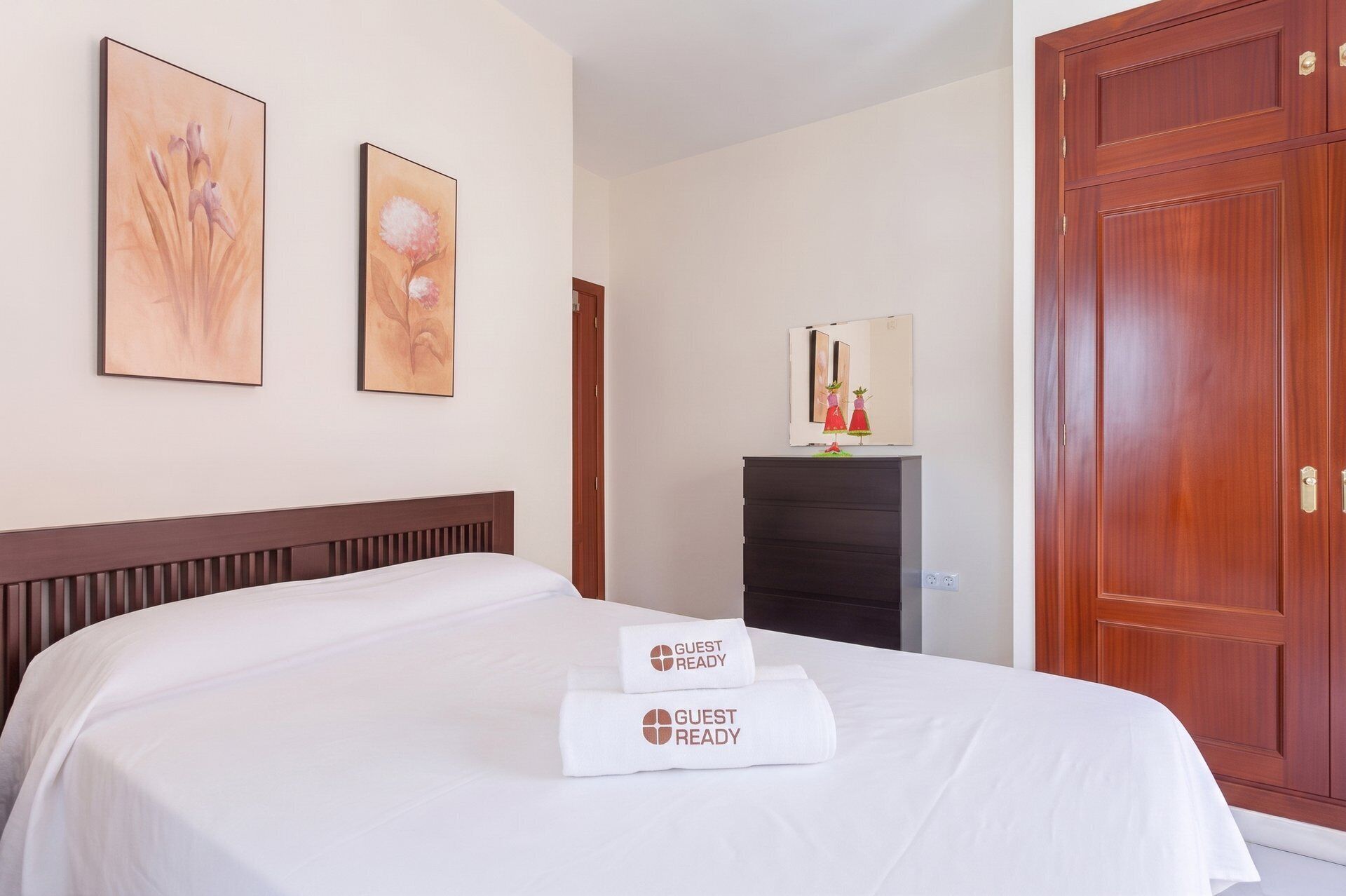 Une chambre d’hôtel comprenant un lit, deux tableaux représentant des motifs floraux encadrés, une commode avec un miroir et une armoire en bois.