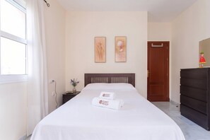 Ein Hotelzimmer mit einem Bett, einem Nachttisch, einer Vase mit Blumen, zwei gerahmten Kunstwerken an der Wand, einer Holztür und einem Schminktisch.