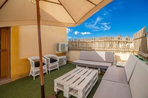 Eine Terrasse mit einer Couch, zwei Korbstühlen, einem Holztisch, einem Sonnenschirm und einer Klimaanlage.