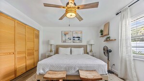 Um quarto com cama, mesas de cabeceira, ventilador de teto, janela com cortinas e um guarda-roupa de madeira.