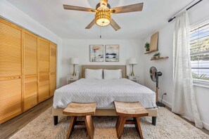 Ein Schlafzimmer mit Bett, Nachttischen, Deckenventilator, einem Fenster mit Vorhängen und einem Holzschrank.