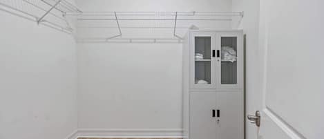 Una habitación con piso de madera, paredes blancas y una estantería metálica de alambre. Se observa un armario blanco con puertas de vidrio.