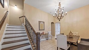 A dining area with a chandelier, a staircase, and a white chair. - Group-Friendly 5BR/4.5BA with Private Yard (Indian Harbour Beach)