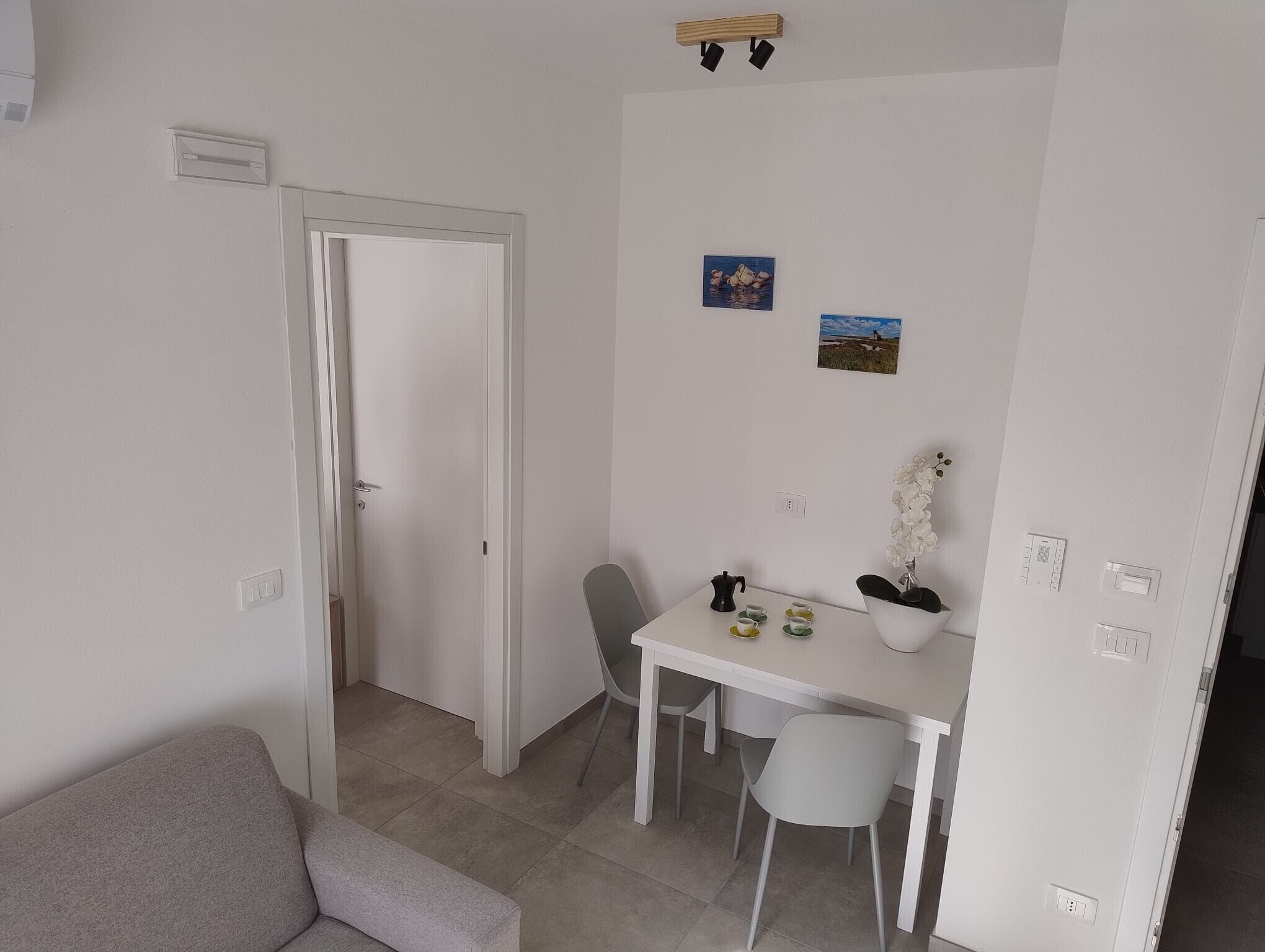 A small, clean room with a white door, a white table with chairs, a grey sofa, and a small potted plant.
