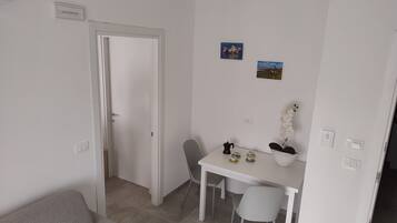 A small, clean room with a white door, a white table with chairs, a grey sofa, and a small potted plant.