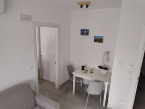 Een kleine, schone kamer met een witte deur, een witte tafel met stoelen, een grijze bank en een kleine bloempot met plant.