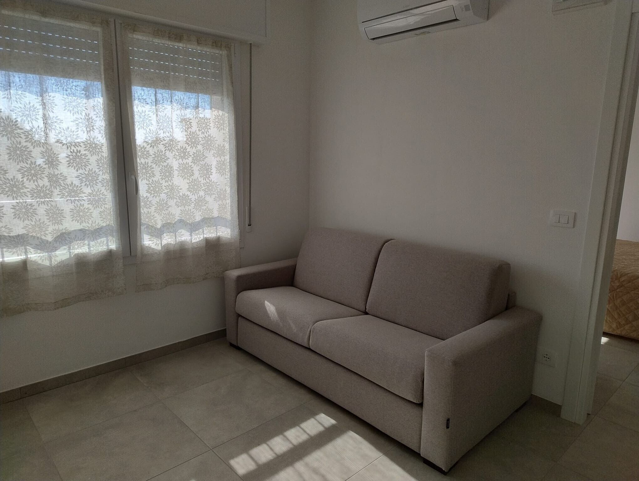 Um quarto com um sofá bege, uma janela com cortinas estampadas e um aparelho de ar condicionado na parede.