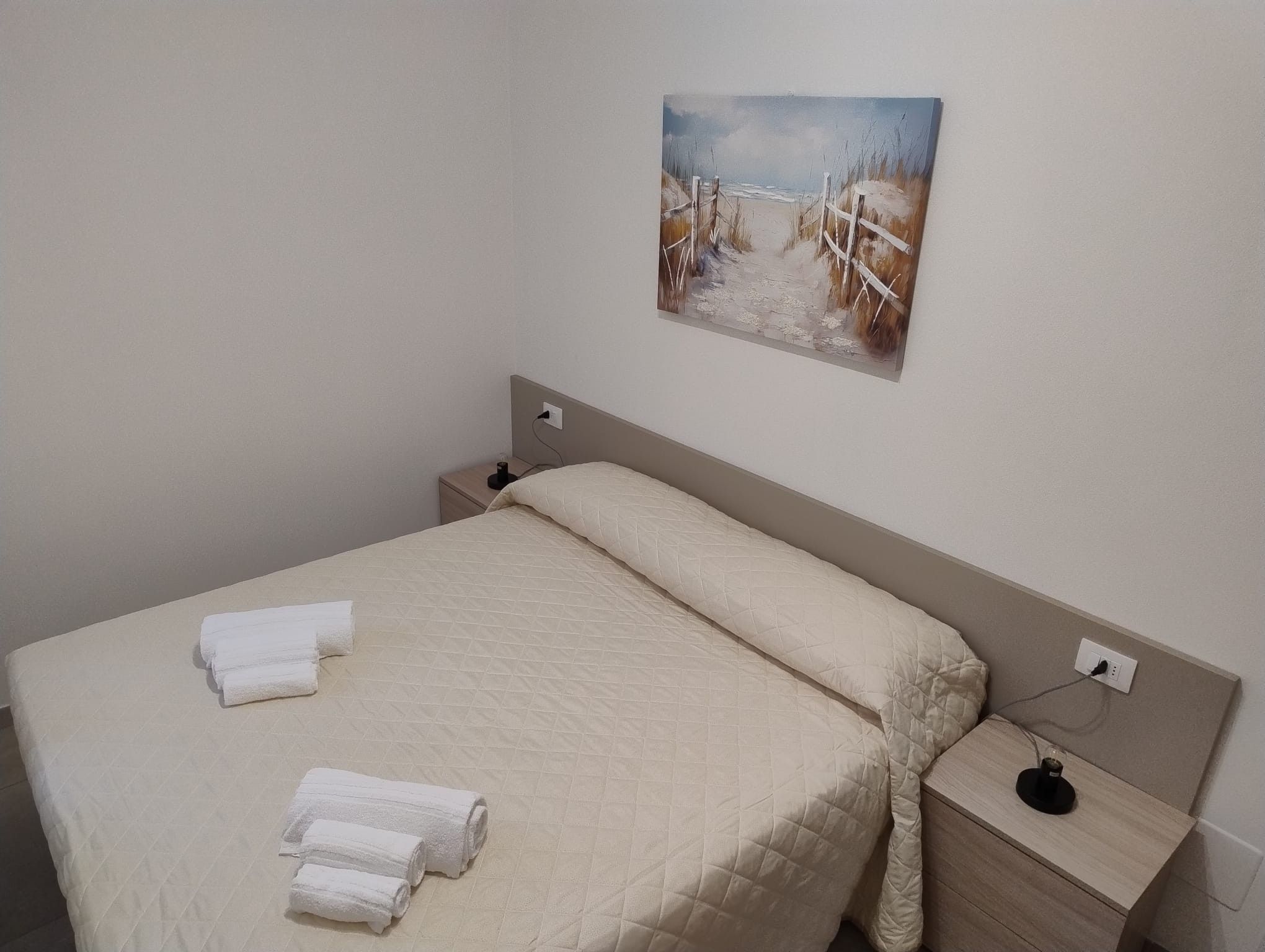 Quarto com cama, mesa de cabeceira, um quadro e tomada na parede.