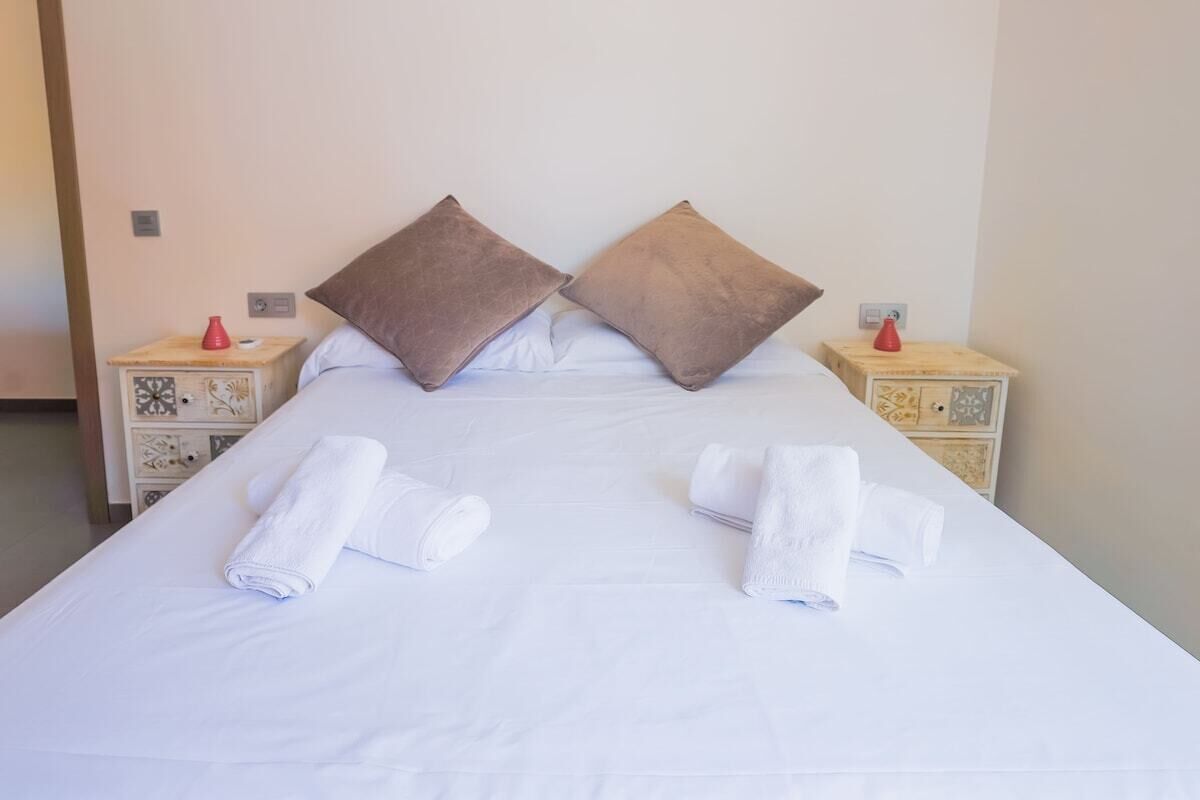 A double bed with white linens, two brown pillows, and two rolled towels on the bed.