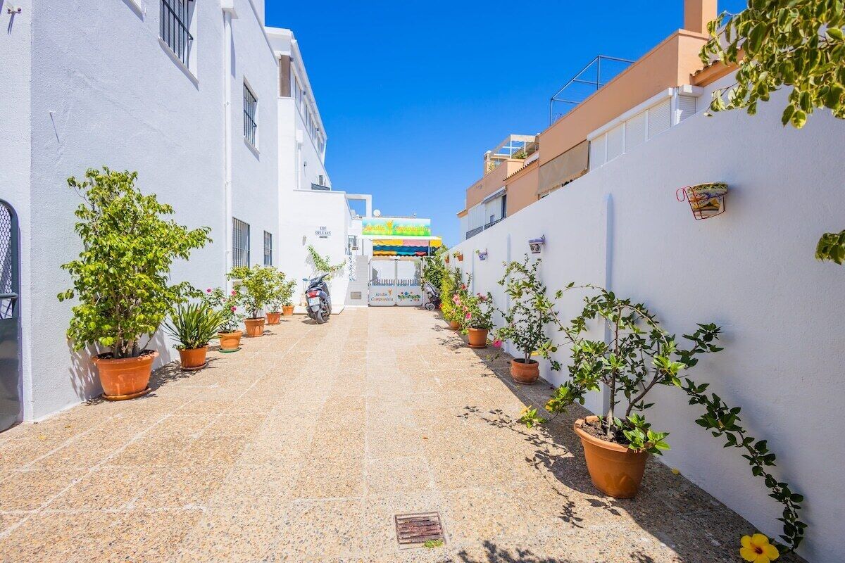 Een smalle straat met potplanten langs de randen, een helderblauwe lucht, en een gebouw met een kleurrijke luifel in de verte.