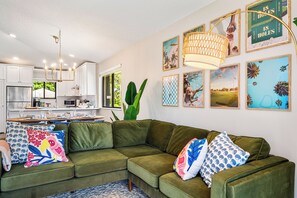 A modern living room with a green sectional sofa, a dining area with a table and chairs, and a kitchen with white cabinets. - Modern 2BR 2BA PGA Home in Paradise, 2 Min to Pool, 15 Min to Beach (Palm Beach Gardens)