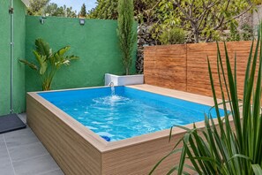 Ein rechteckiges Whirlpool mit Holzrahmen und blauem Innenraum, umgeben von einer grünen Wand und einem Holzzaun, mit Pflanzen und Bäumen im Hintergrund.