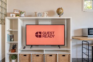 Ein Fernseher an einer weißen Unterhaltungseinheit, mit einem 'GUEST READY' Schild, umgeben von Regalen mit Büchern, Dekorationsgegenständen und Korbwannen.