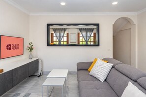A modern living room with a grey sofa, a white table, and a framed artwork above a window with curtains. - GuestReady - Urban Suite City Center (La Laguna)