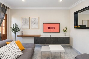 Ein modernes Wohnzimmer mit einem grauen Sofa, einem weißen Couchtisch und einem an der Wand montierten Flachbildfernseher. Darüber befinden sich zwei gerahmte Stadtansichten, darunter ein kleines Zimmerpflänzchen auf einem Regal.