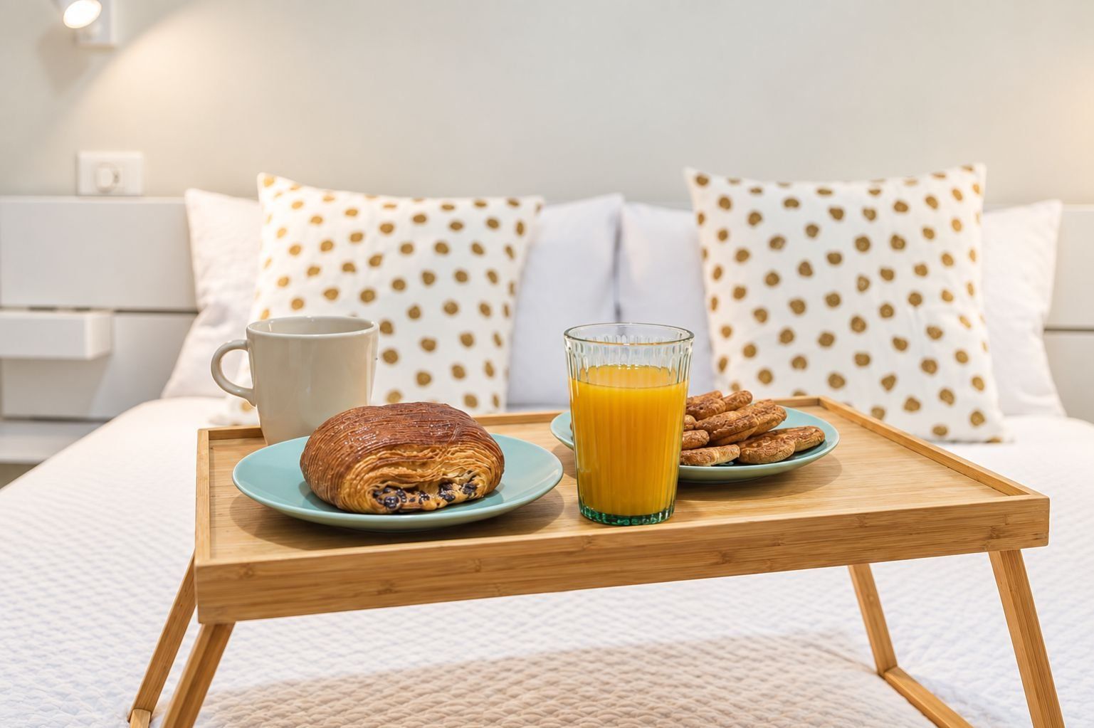 Een ontbijfwagen met gebak, een glas sinaasappelsap en koekjes op een bed met wit beddegoed en stippelhoeden kussens.