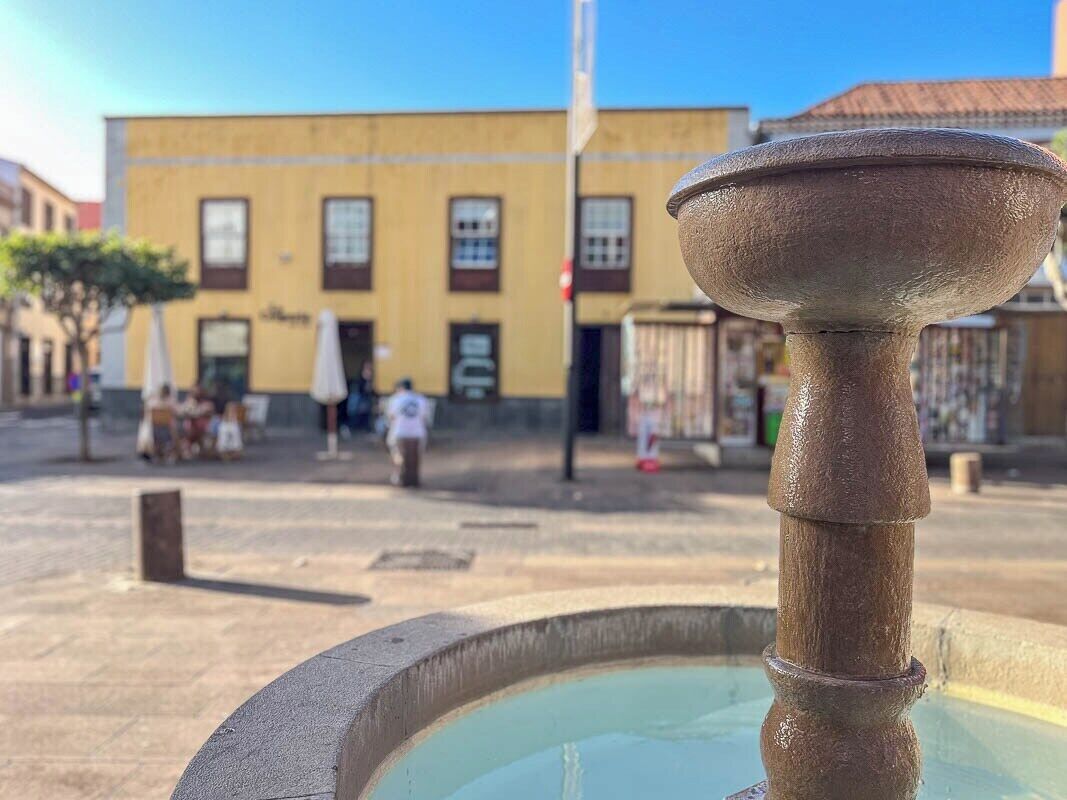 Une fontaine dotée d’une colonne centrale et d’un bassin circulaire, située devant un bâtiment aux nombreuses fenêtres, avec un ciel dégagé en arrière-plan.