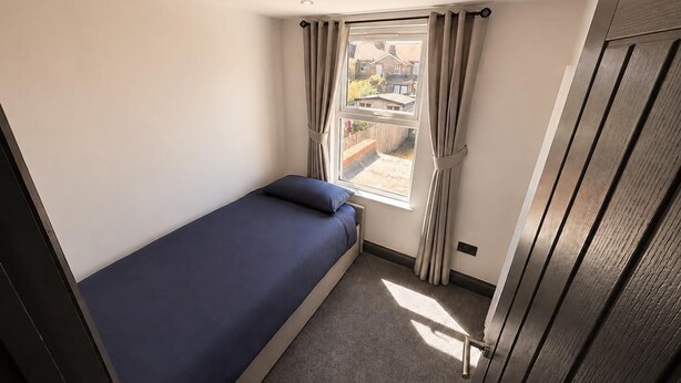 Ein kleines Zimmer mit einem Einzelbett, einem Fenster, Vorhängen und Blick ins Freie.