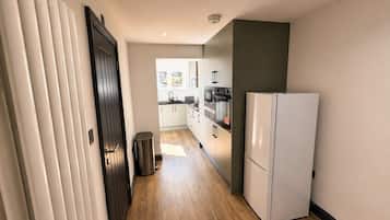 A narrow hallway with a wooden floor, a white refrigerator, a trash bin, and a door leading to a kitchen area with cabinets and appliances.