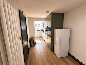 A narrow hallway with a wooden floor, a white refrigerator, a trash bin, and a door leading to a kitchen area with cabinets and appliances.