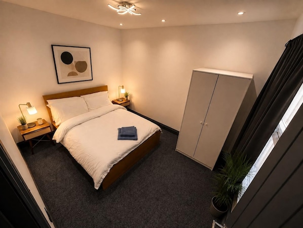 A hotel room with a bed, bedside tables, a wardrobe, a framed artwork, and a ceiling light fixture.