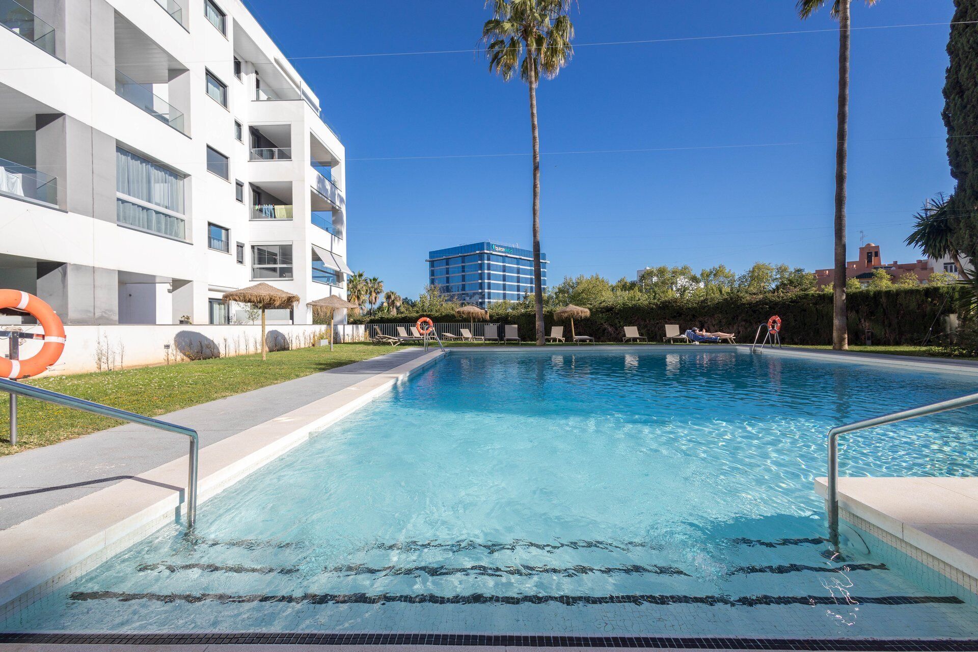 A swimming pool with a clear blue water surface, surrounded by a white building with balconies, a palm tree, and a lifebuoy.